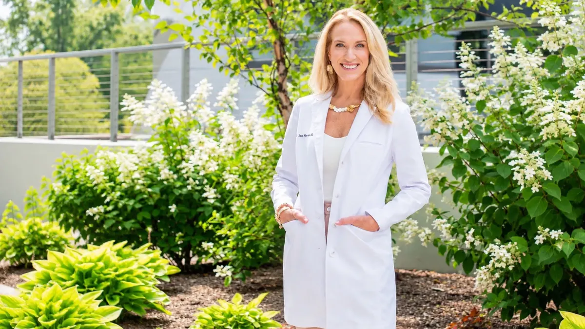 Doctor standing outdoors in a white coat, smiling, with greenery and blooming flowers in the background.