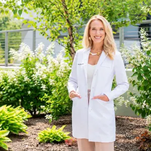 Doctor standing outdoors in a white coat, smiling, with greenery and blooming flowers in the background.