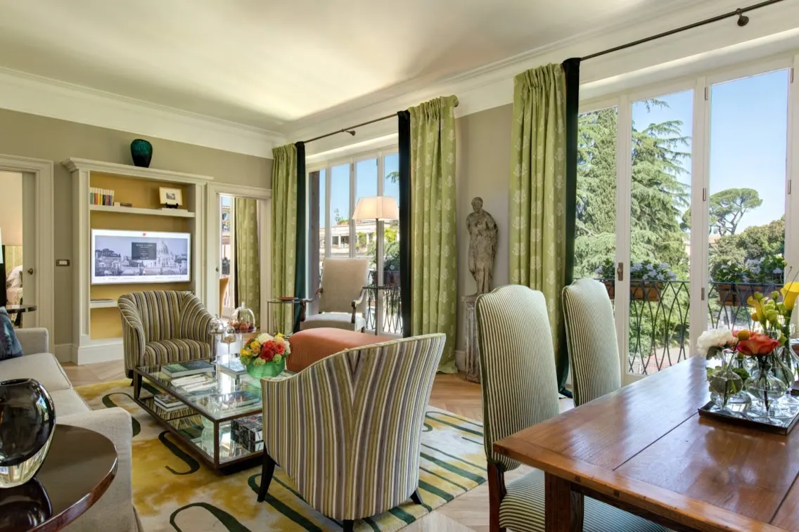 Elegant suite interior featuring striped chairs, a wooden dining table, and large windows with green curtains overlooking a garden.