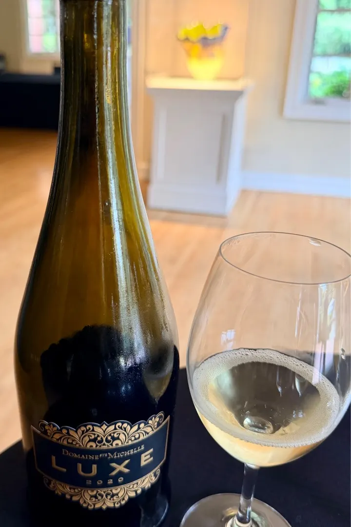 A bottle of Domaine Ste. Michelle Luxe sparkling wine next to a filled glass, set in a bright, inviting interior.