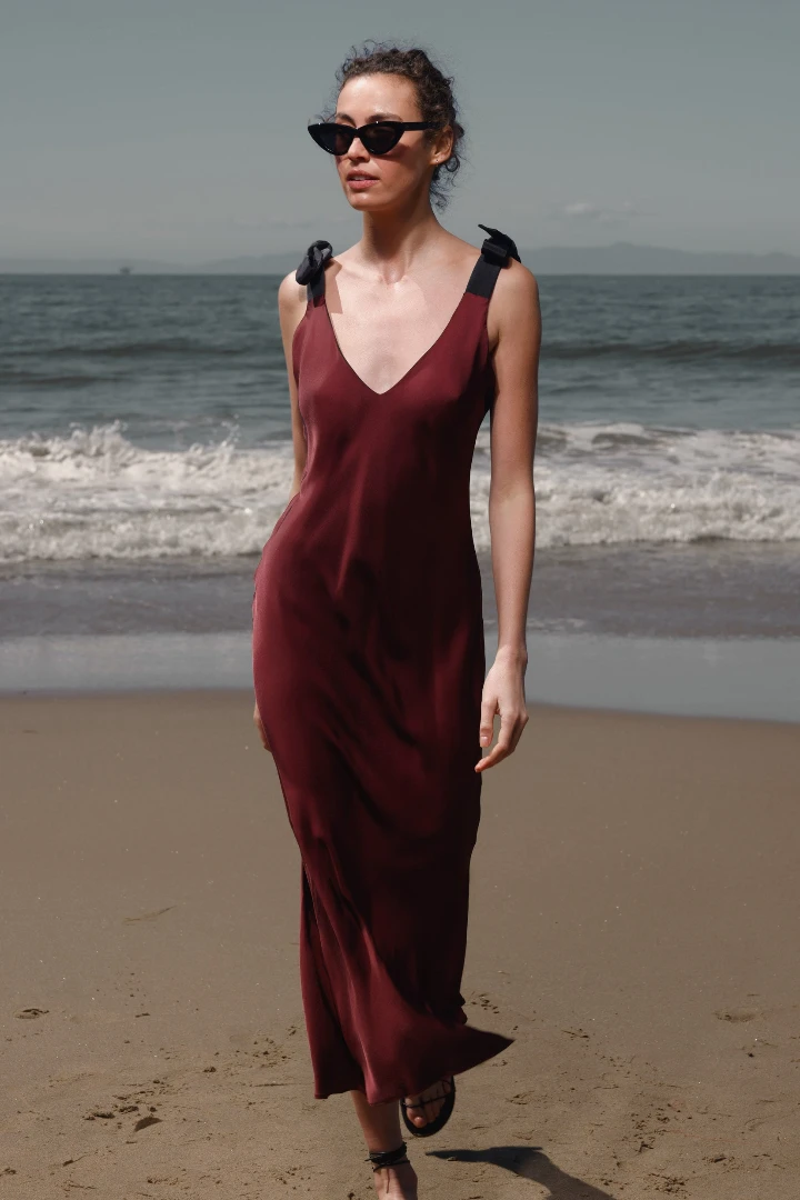 Model walking along the beach in a burgundy dress with shoulder knots, paired with black sunglasses and sandals.