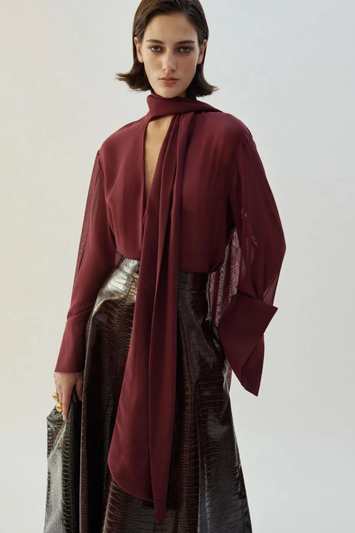 Model wearing a deep burgundy top with a scarf detail and a shiny black skirt, showcasing a stylish fall outfit.