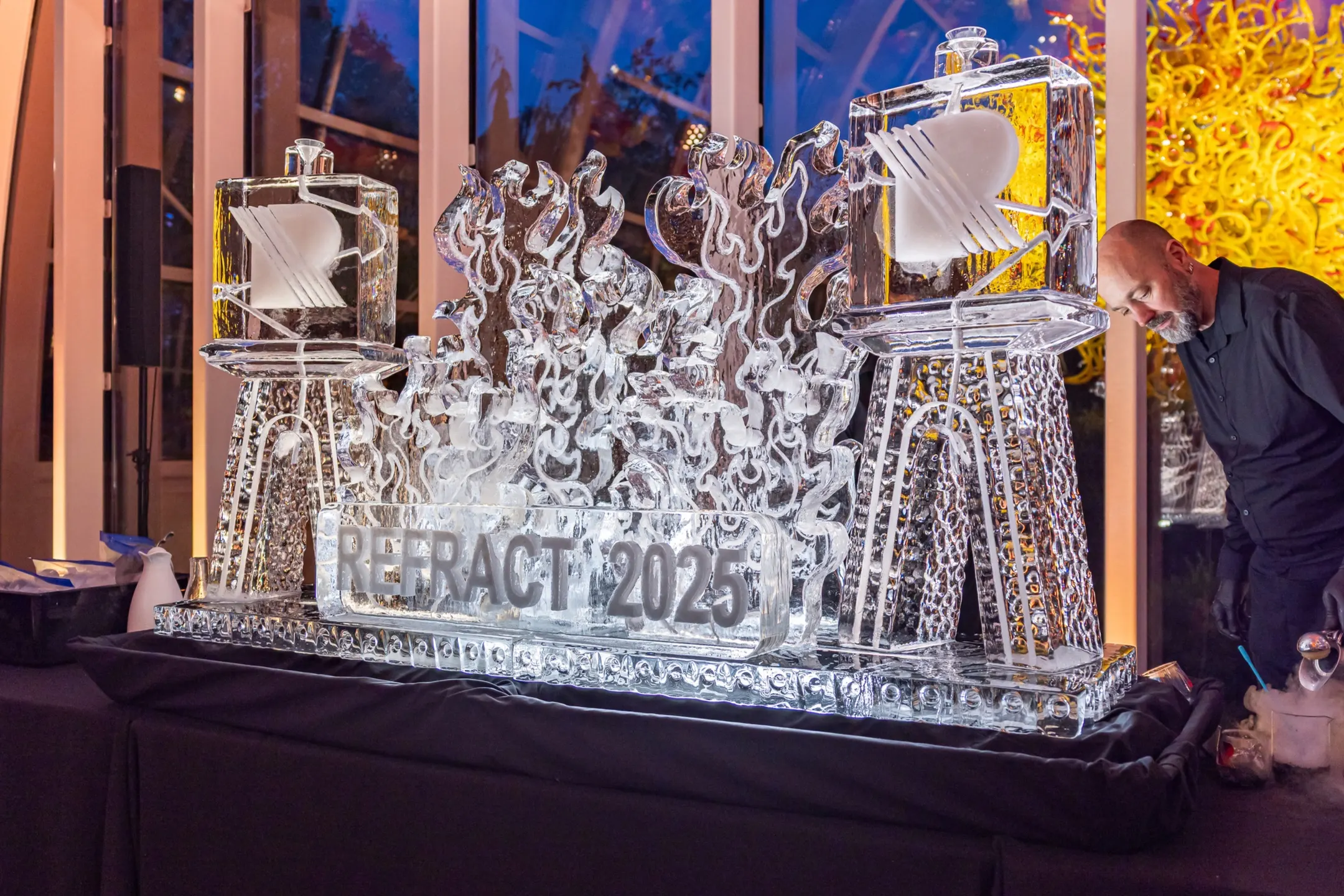 Intricate ice sculpture displaying "REFRACT 2025" with stylized flames, set up at Chihuly Garden and Glass for the opening night event.