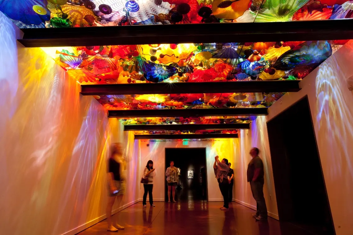 Colorful glass art hangs from the ceiling in a gallery, with several visitors exploring the vibrant space beneath.