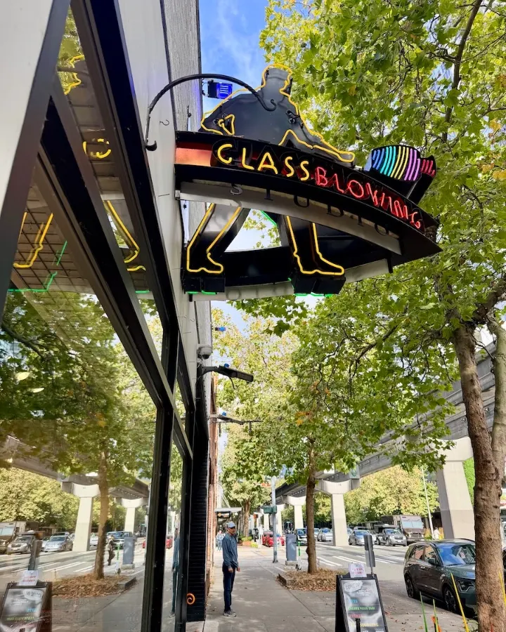Neon sign for a glassblowing studio in Seattle, surrounded by trees and city streets, reflecting an artistic atmosphere.