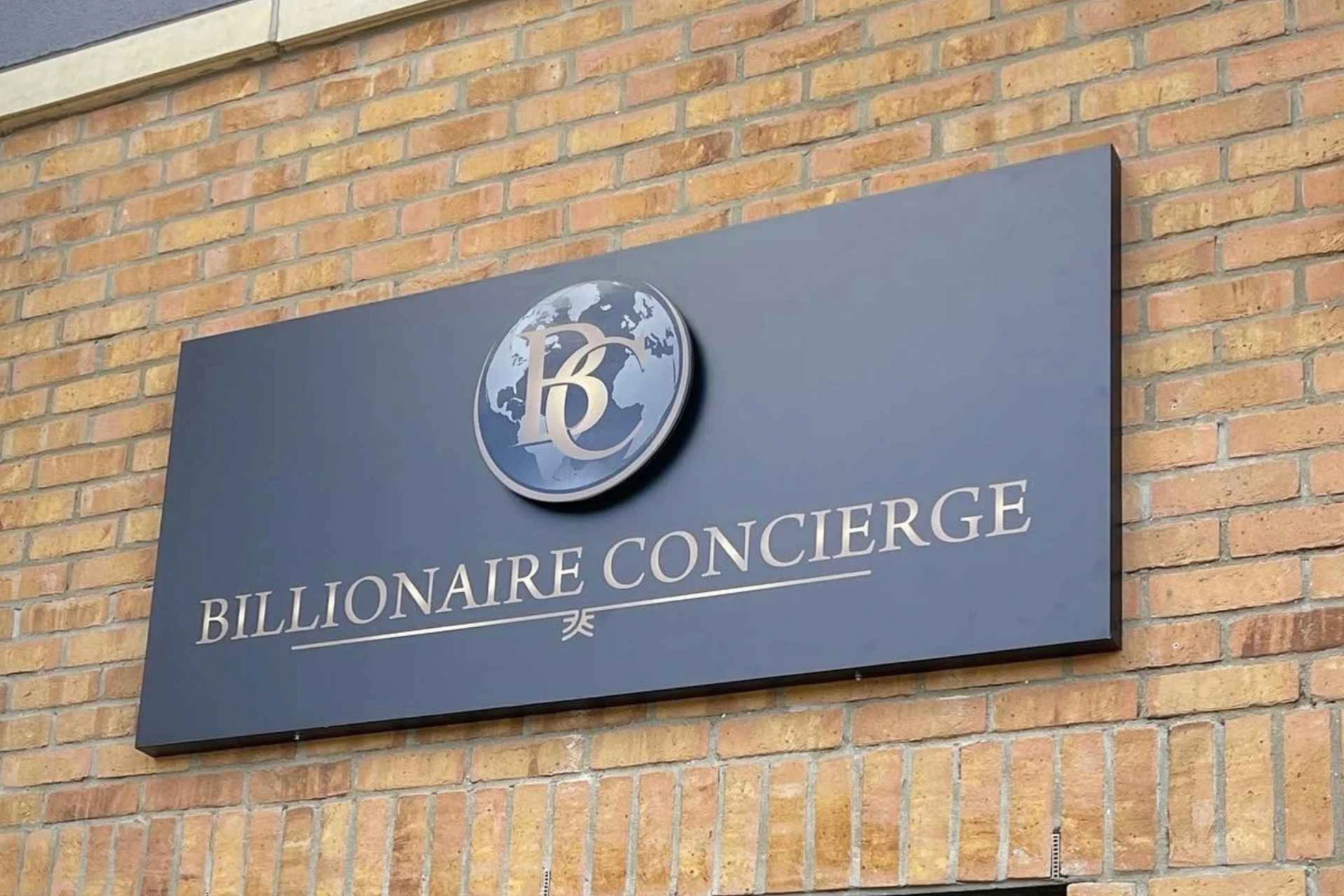 Billionaire Concierge sign on a brick wall, featuring a logo with a globe and the company name in elegant lettering.