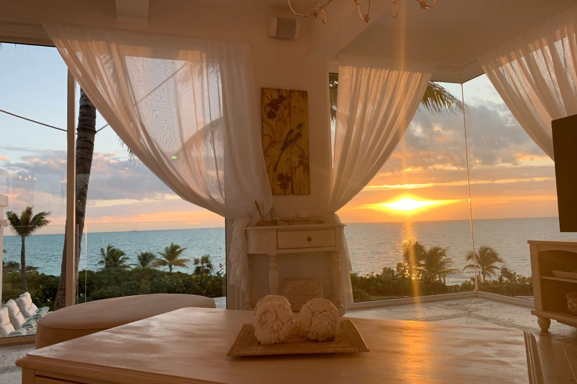 Sunset view from a luxury interior with sheer curtains, palms lining the coastline, and a serene ocean backdrop.