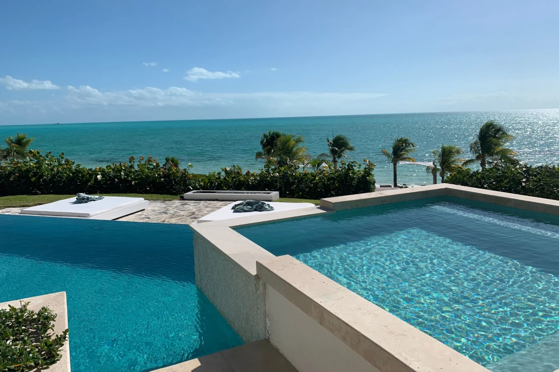 Luxurious infinity pool overlooking the ocean, surrounded by palm trees and sun loungers on a bright, sunny day.