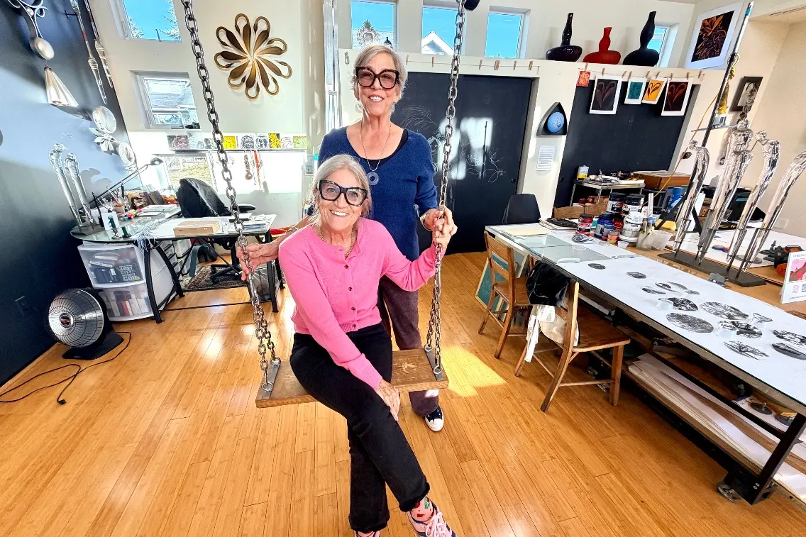 Two women posed on a swing in an art studio, surrounded by creative materials and colorful artworks.