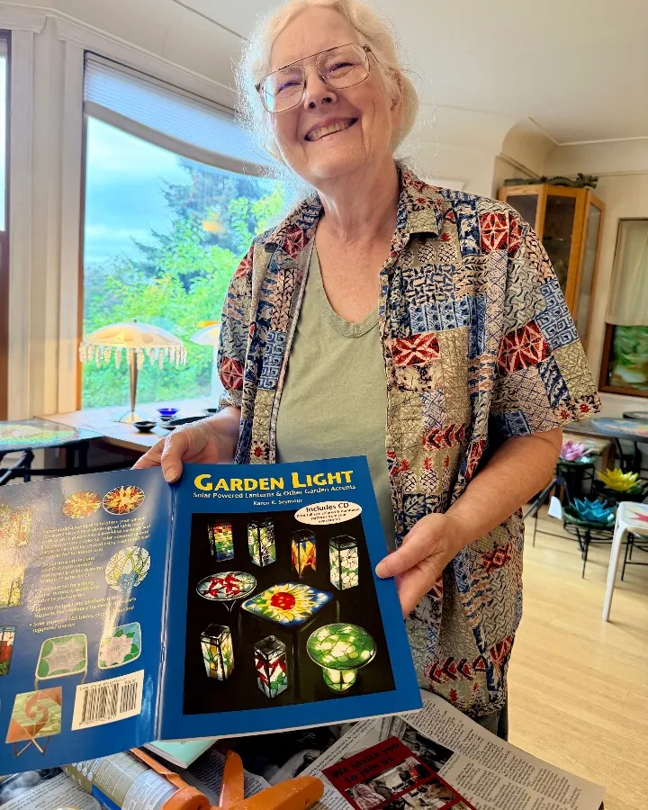 A woman smiles while holding a book titled "Garden Light," featuring colorful garden accents in a bright, cozy studio.