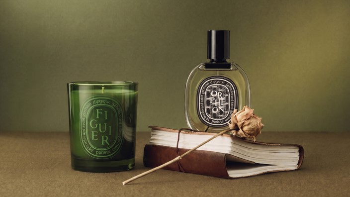 A green candle labeled "Figuer" beside a perfume bottle and a dried rose on a brown book with a textured cover.