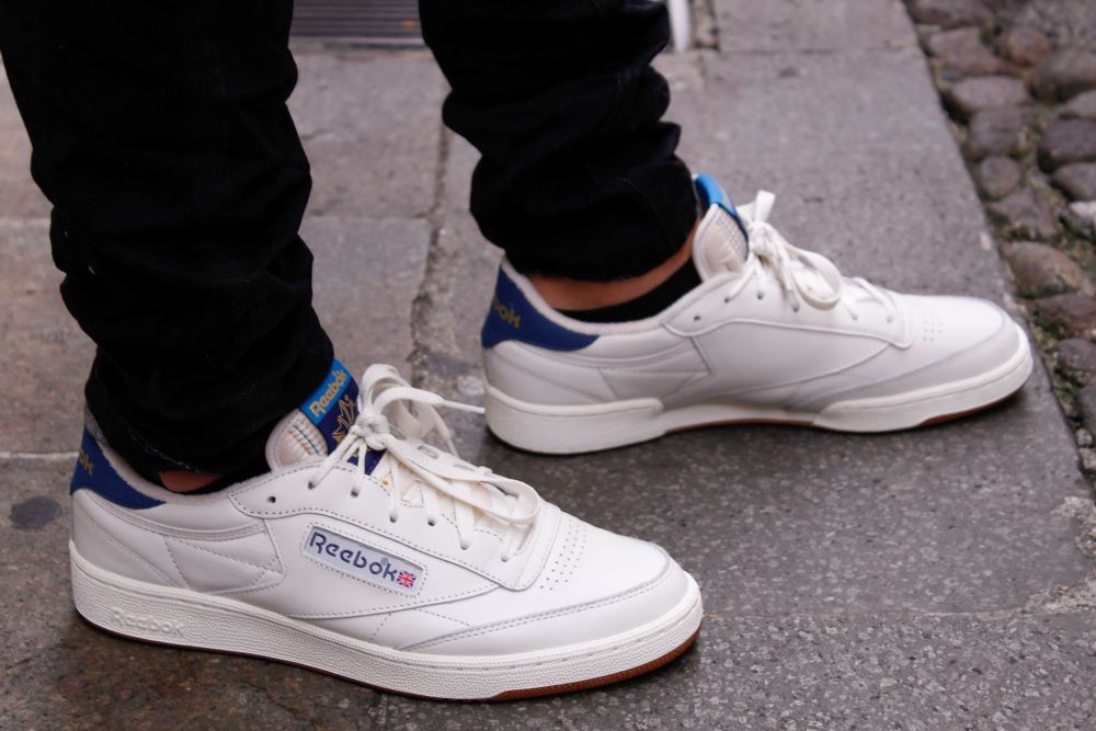 Casual white Reebok trainers worn with black jeans on a stone pavement.