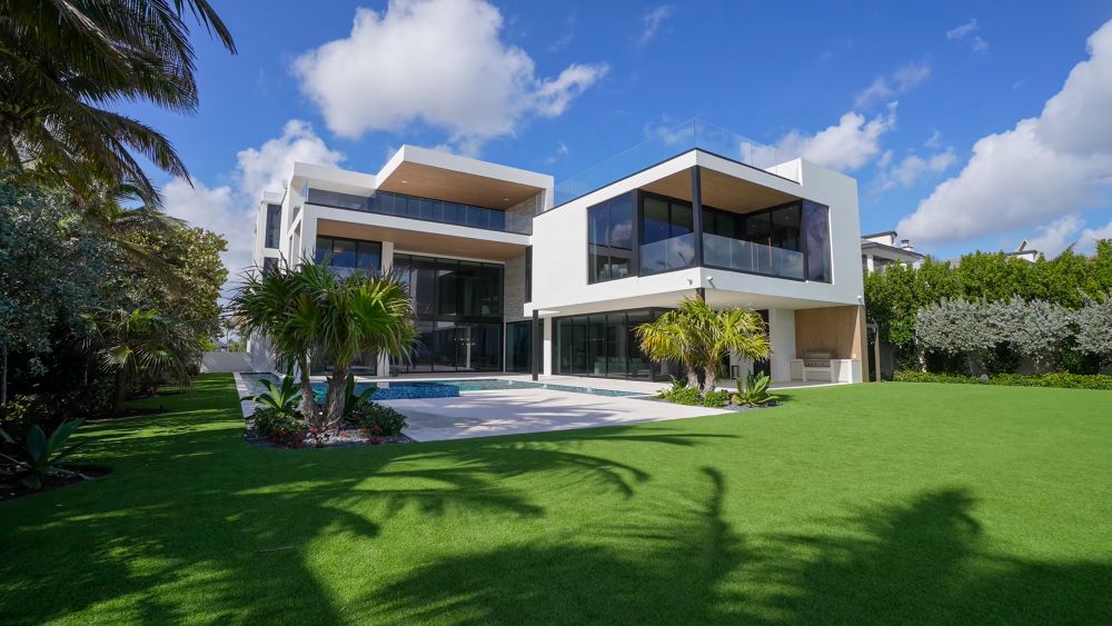 Modern luxury home with large glass windows, a spacious lawn, and tropical landscaping under a blue sky.