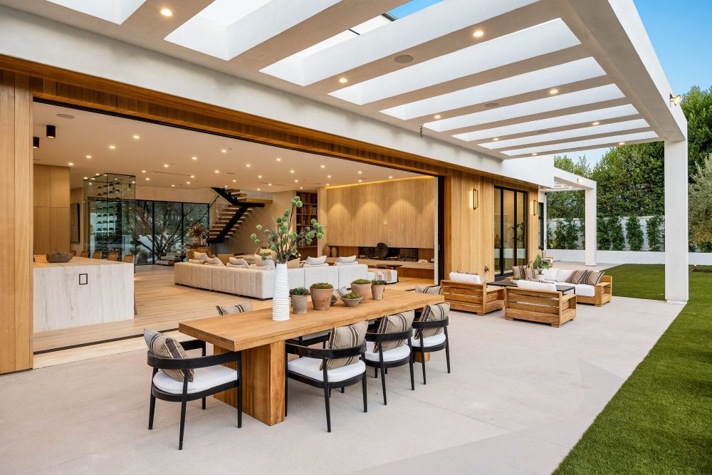 Modern outdoor living space with wooden dining table, stylish seating area, and large glass doors opening to a bright interior.