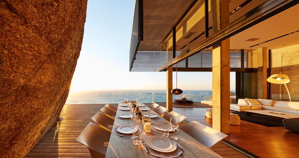 Modern dining area with a long table set for dinner, large windows offering a view of the ocean, and a warm sunset ambiance.