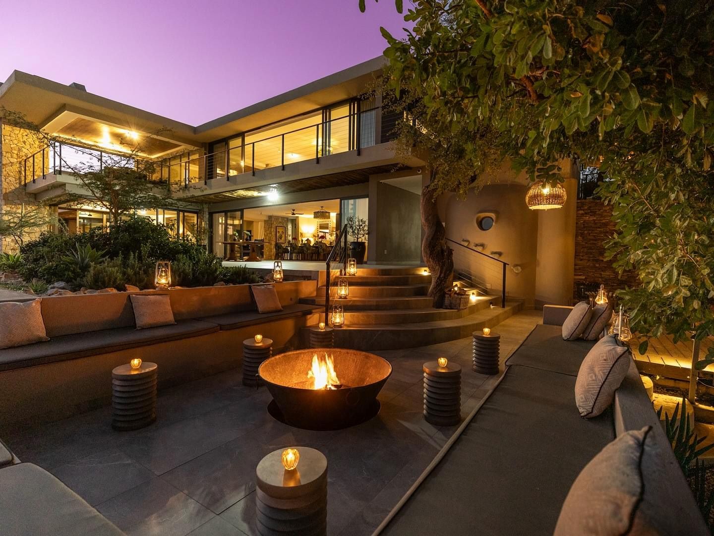 Modern outdoor seating area with a fire pit, surrounded by lush greenery and illuminated by lanterns against a twilight sky.