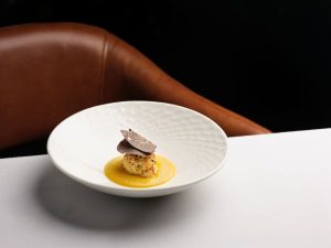 Truffle-topped dish served with a bright yellow sauce in a white bowl on a table, with a brown chair in the background.