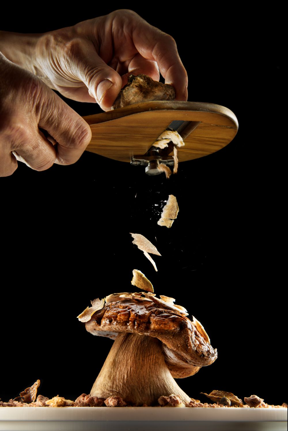 Hands grating a truffle over a porcini mushroom, with shavings falling onto the dish against a black background.