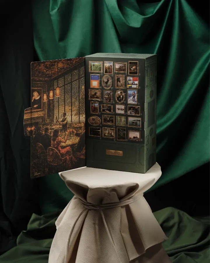 A decorative whiskey gift box with images and scenes displayed, resting on a pedestal against a green backdrop.