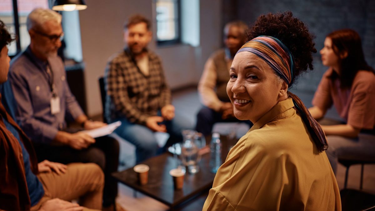 A woman smiles while seated in a support group session surrounded by other participants.
