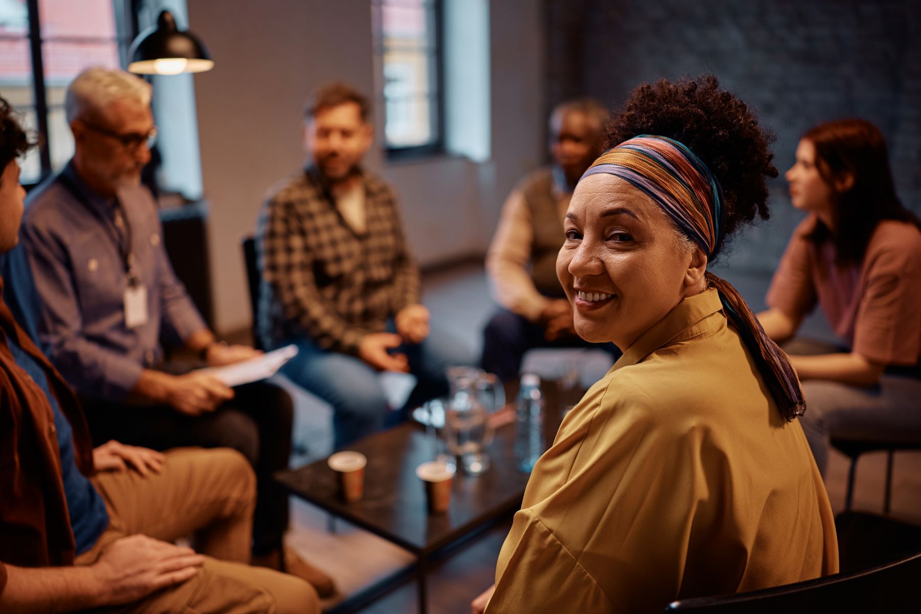 A woman smiles while seated in a support group session surrounded by other participants.