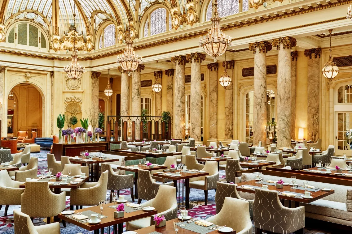 Elegant dining area in The Garden Court at the Palace Hotel, featuring chandeliers, marble columns, and tables set for guests.