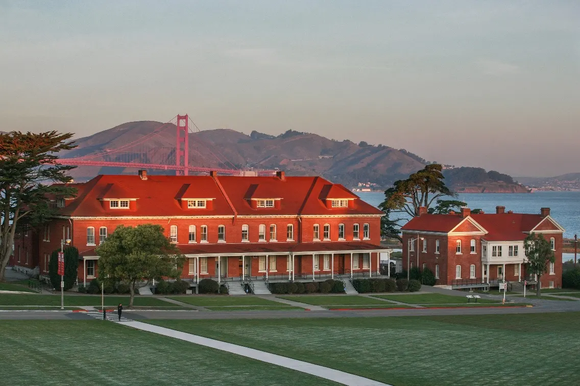 Historic red-brick buildings of the Lodge at Presidio with the Golden Gate Bridge in the background at sunset.