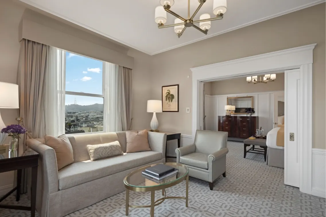 Luxurious hotel suite with a view of San Francisco, featuring a cozy sofa, armchair, and stylish decor.