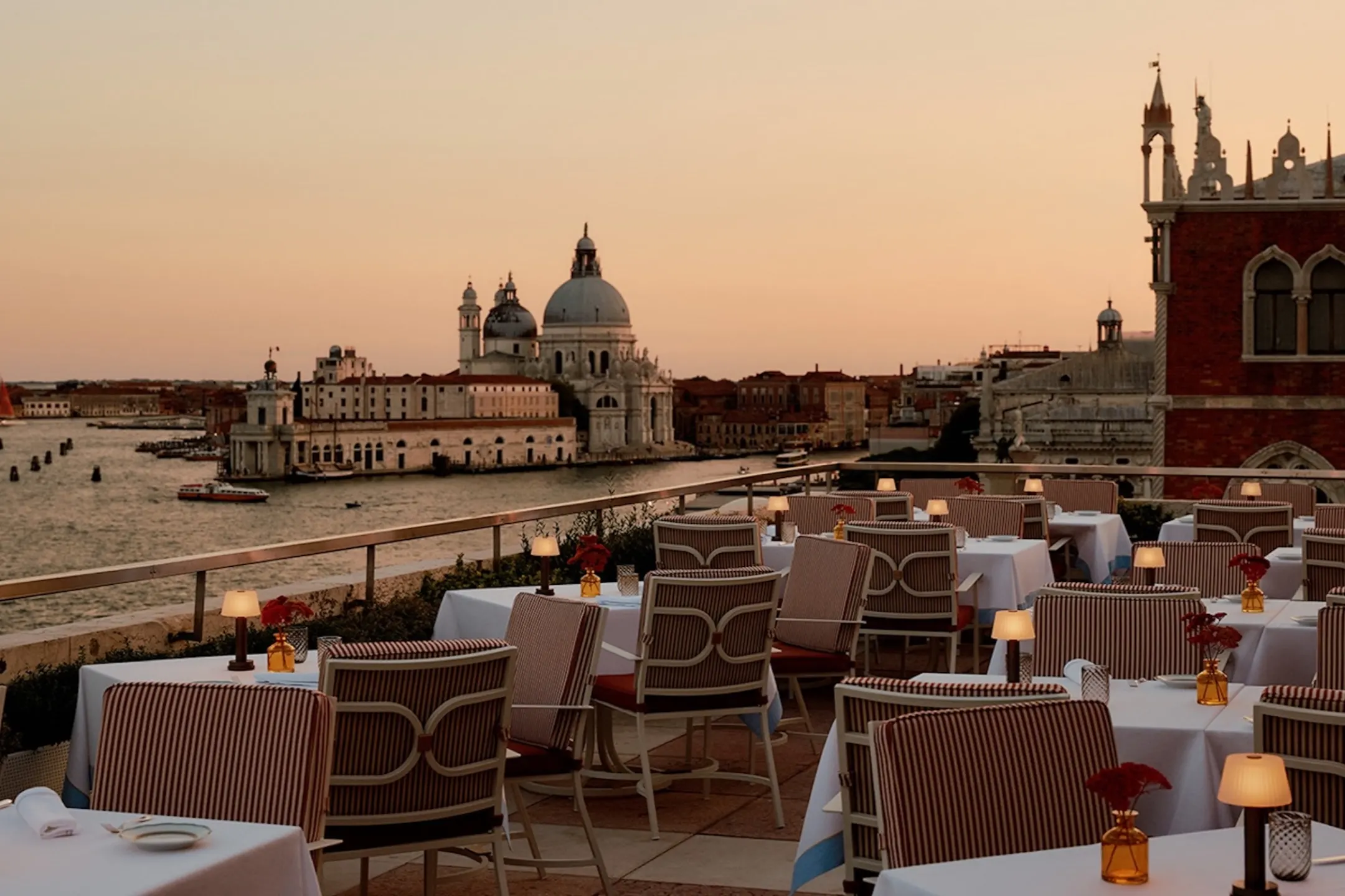 Dining tables set on a terrace with a view of Venice's stunning architecture and bustling waterways at sunset.
