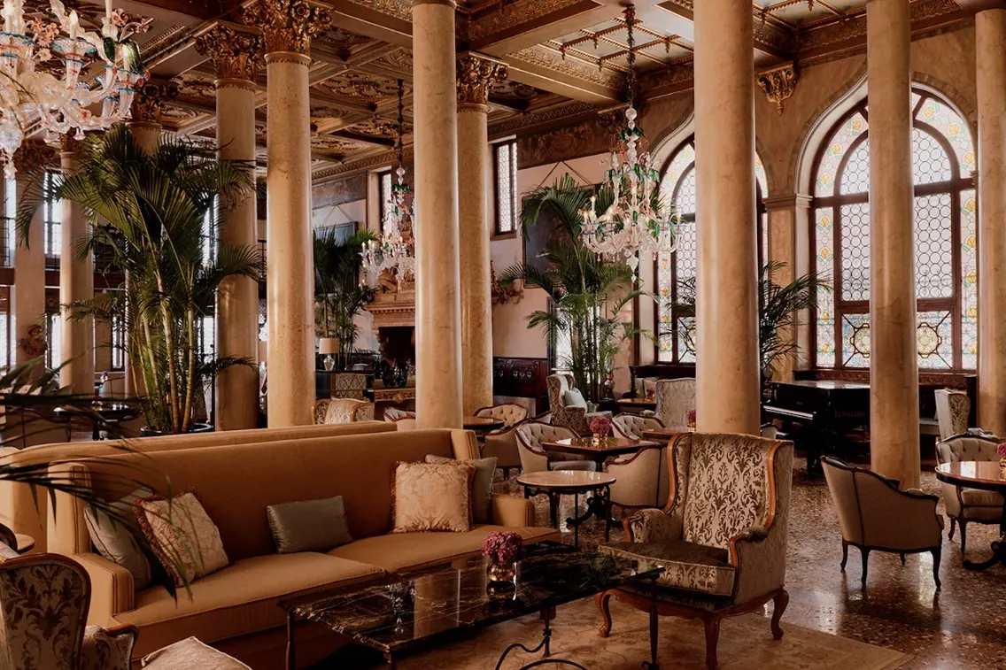 Luxurious hotel lobby featuring elegant furnishings, ornate chandeliers, and palm plants in a grand Venetian palazzo.