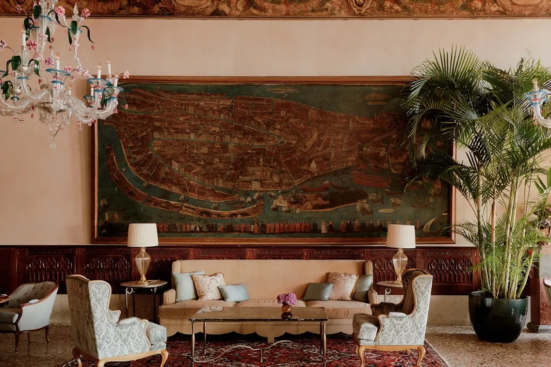 Luxurious hotel lobby featuring elegant seating, a large historical painting of Venice, and lush greenery.