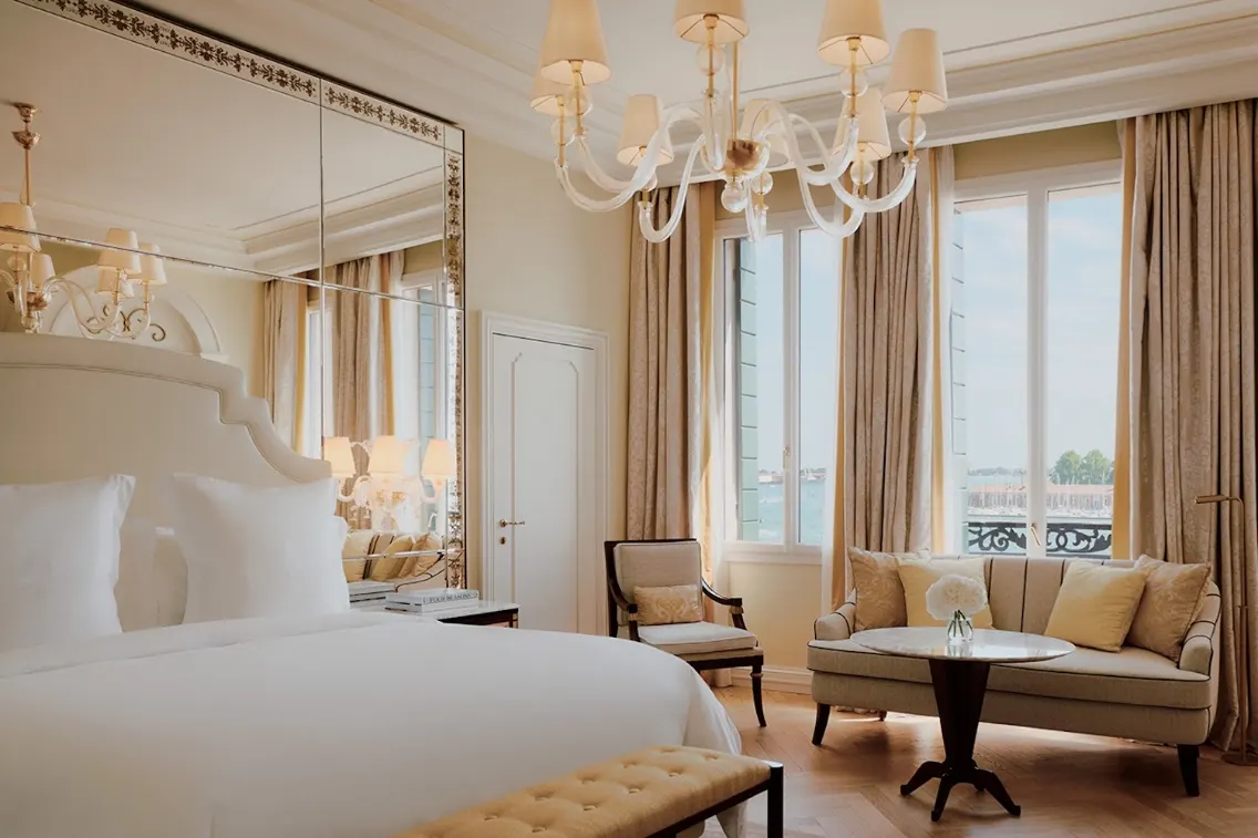 Luxurious hotel room featuring a plush bed, elegant furnishings, large windows with natural light, and a stunning chandelier.
