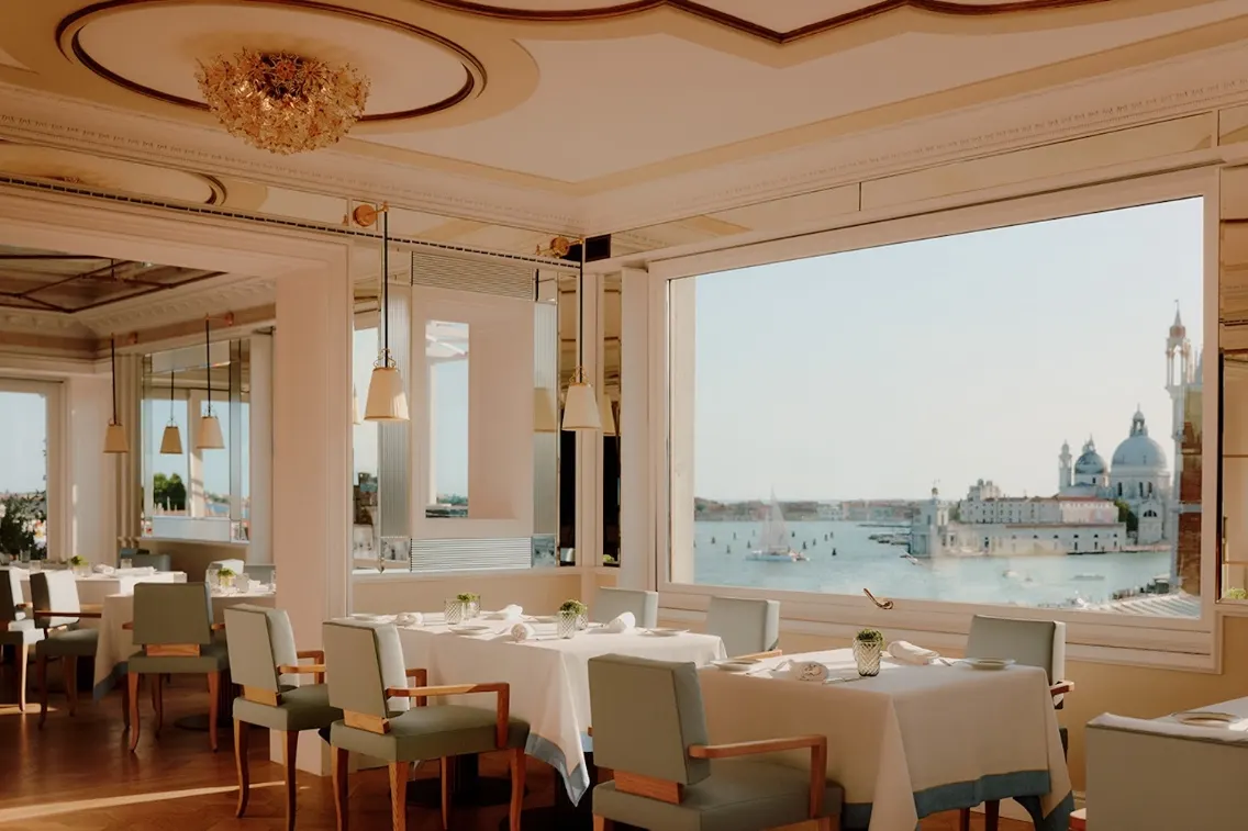 Elegant dining area with tables and chairs, showcasing stunning views of the Venice waterfront and historic architecture.