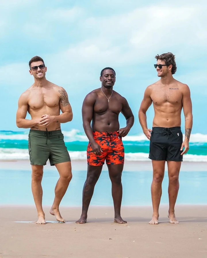 Three men in swim trunks stand on a beach with ocean waves in the background, showcasing a variety of swimwear styles.