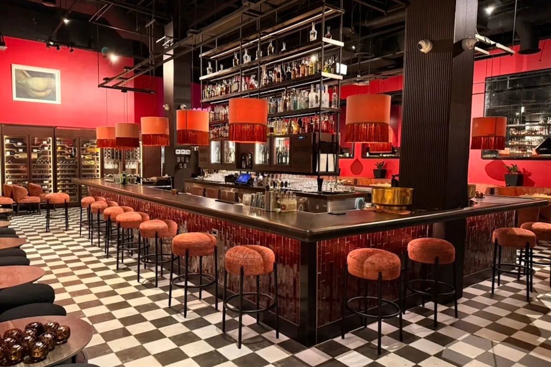 Trendy bar interior featuring a checkered floor, vibrant red walls, overhead orange lamps, and stylish seating at the counter.