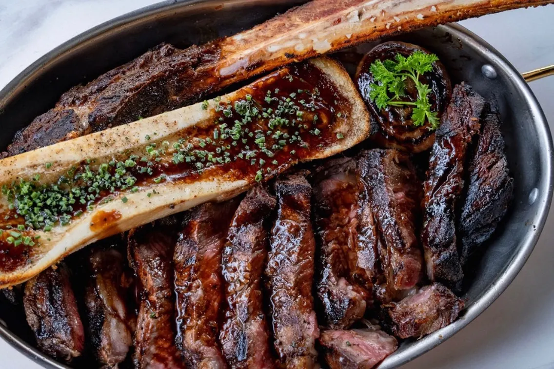 A platter of grilled meats including prime rib, bone marrow, and garnished with herbs, served in a rustic metal dish.