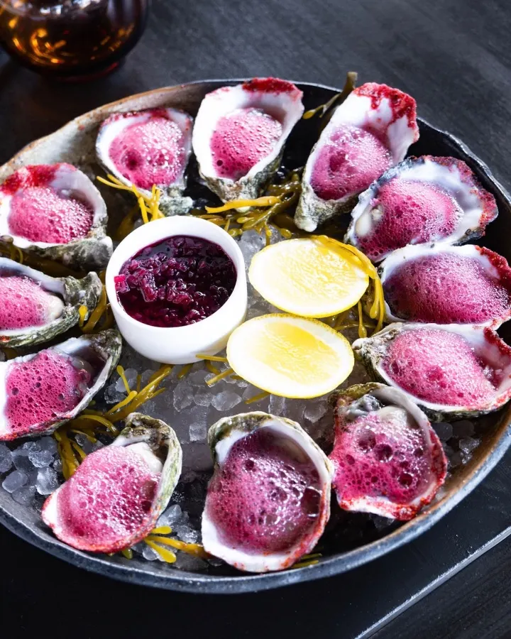 Oysters topped with a berry foam and served with lemon halves and a side of berry sauce on a plate of ice.