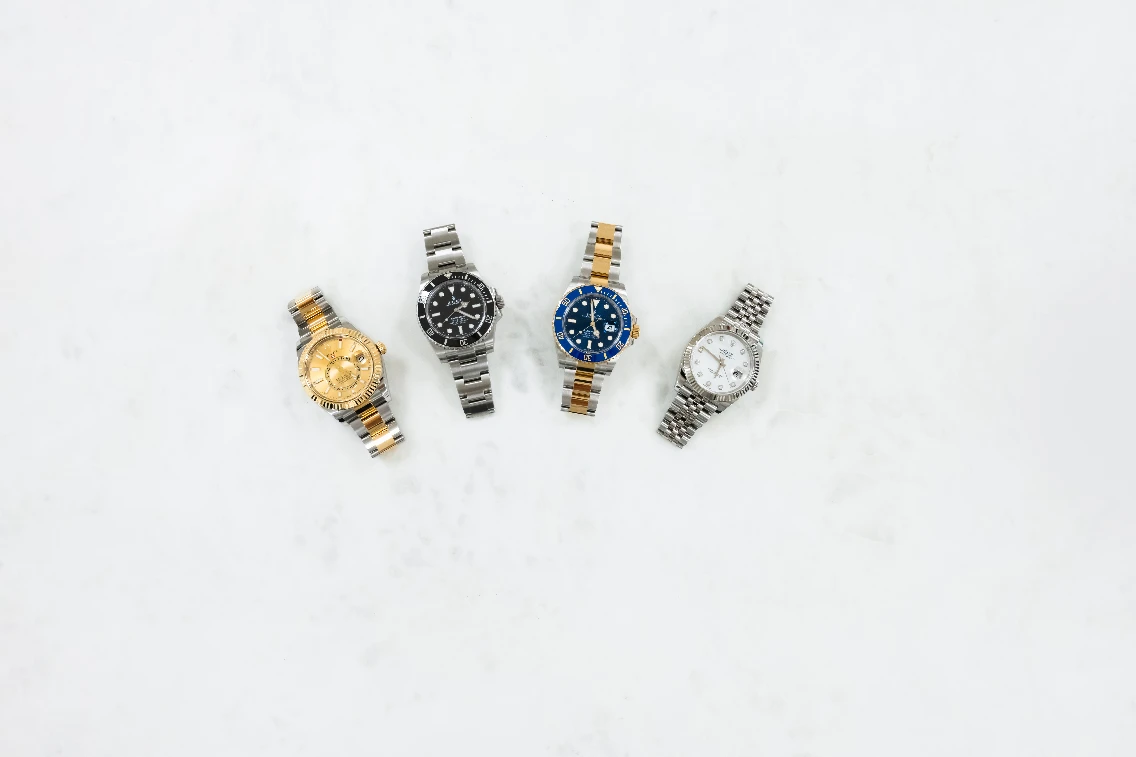Four luxury Rolex watches displayed on a light marble surface, featuring different styles and colors.