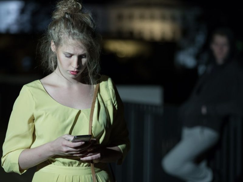 A woman in a yellow dress looks intently at her smartphone while a figure looms in the background, highlighting safety concerns.
