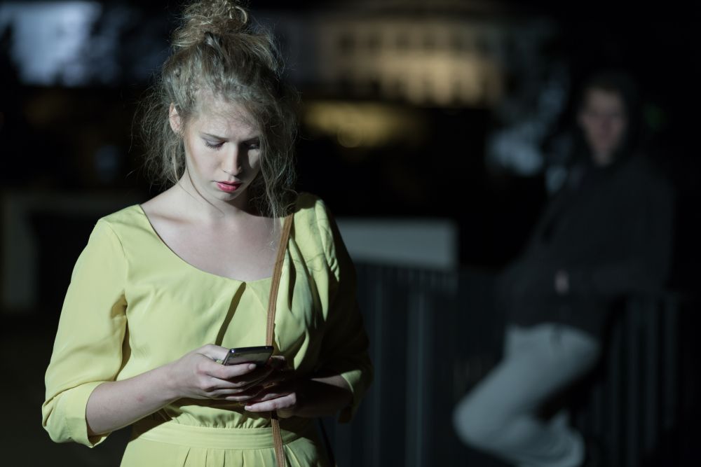 A woman in a yellow dress looks intently at her smartphone while a figure looms in the background, highlighting safety concerns.