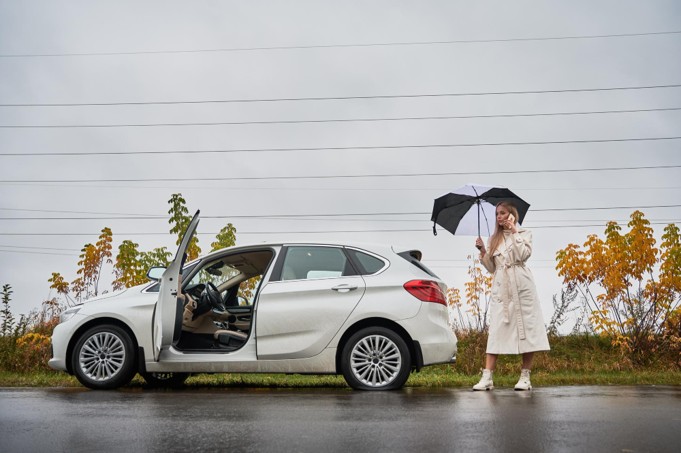 A woman holding an umbrella stands next to an open-door luxury car on a rainy day surrounded by autumn foliage.