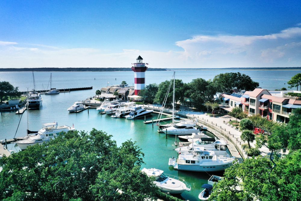 Waterfront view of a marina with yachts, a striped lighthouse, and vibrant buildings on Hilton Head Island.