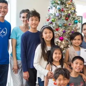 A group of smiling children and adults gathered around a decorated Christmas tree, holding a sign that says "Because I'm a Miracle!"