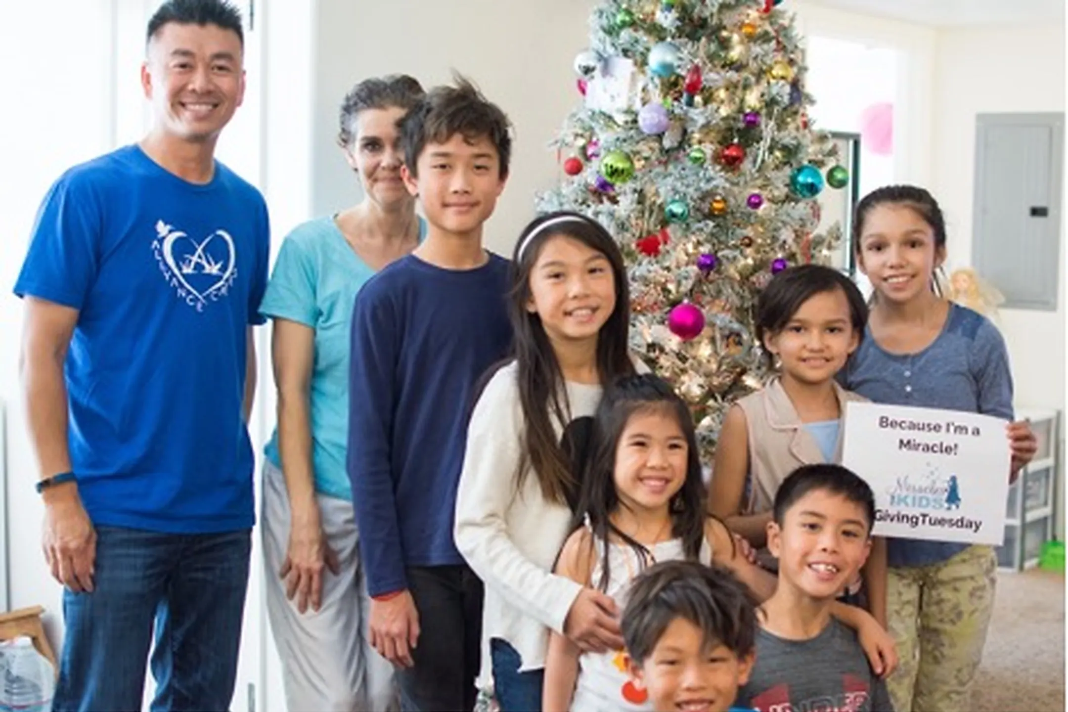 A group of smiling children and adults gathered around a decorated Christmas tree, holding a sign that says "Because I'm a Miracle!"