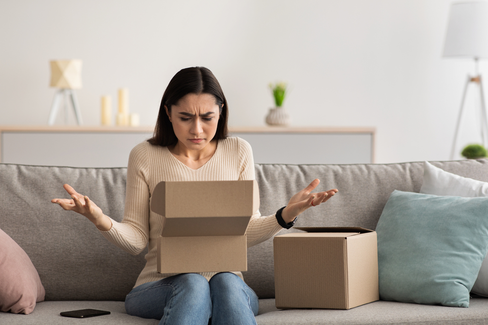 Frustrated woman opening a small box on a couch, expressing disappointment over the contents.
