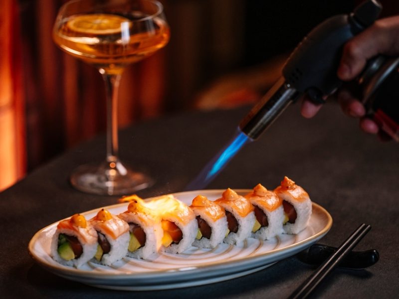 Sushi rolls being seared with a kitchen torch, accompanied by a cocktail on a table.