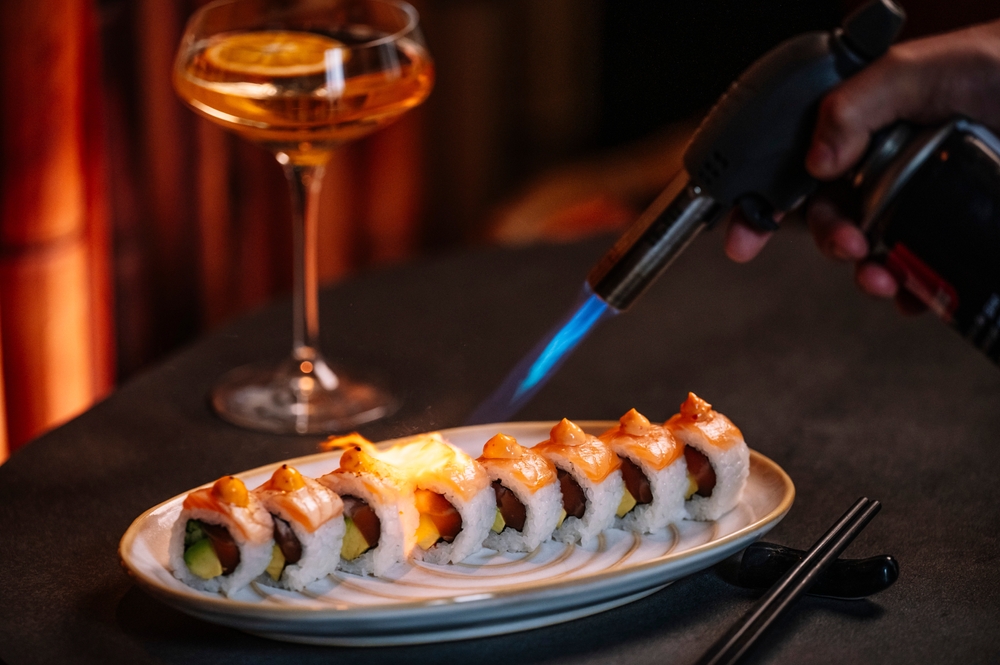 Sushi rolls being seared with a kitchen torch, accompanied by a cocktail on a table.