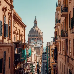 Narrow street in Malta with historic buildings and a prominent dome in the background.