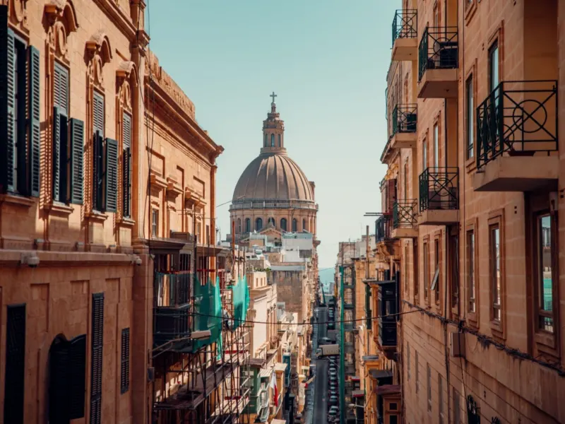 Narrow street in Malta with historic buildings and a prominent dome in the background.