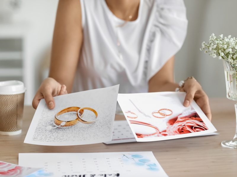 A person examines printed images of wedding rings and floral arrangements while seated at a table with a coffee cup.